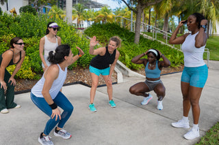 Group of women exercising outdoors on a sidewalk with greenery and palm trees in the background.